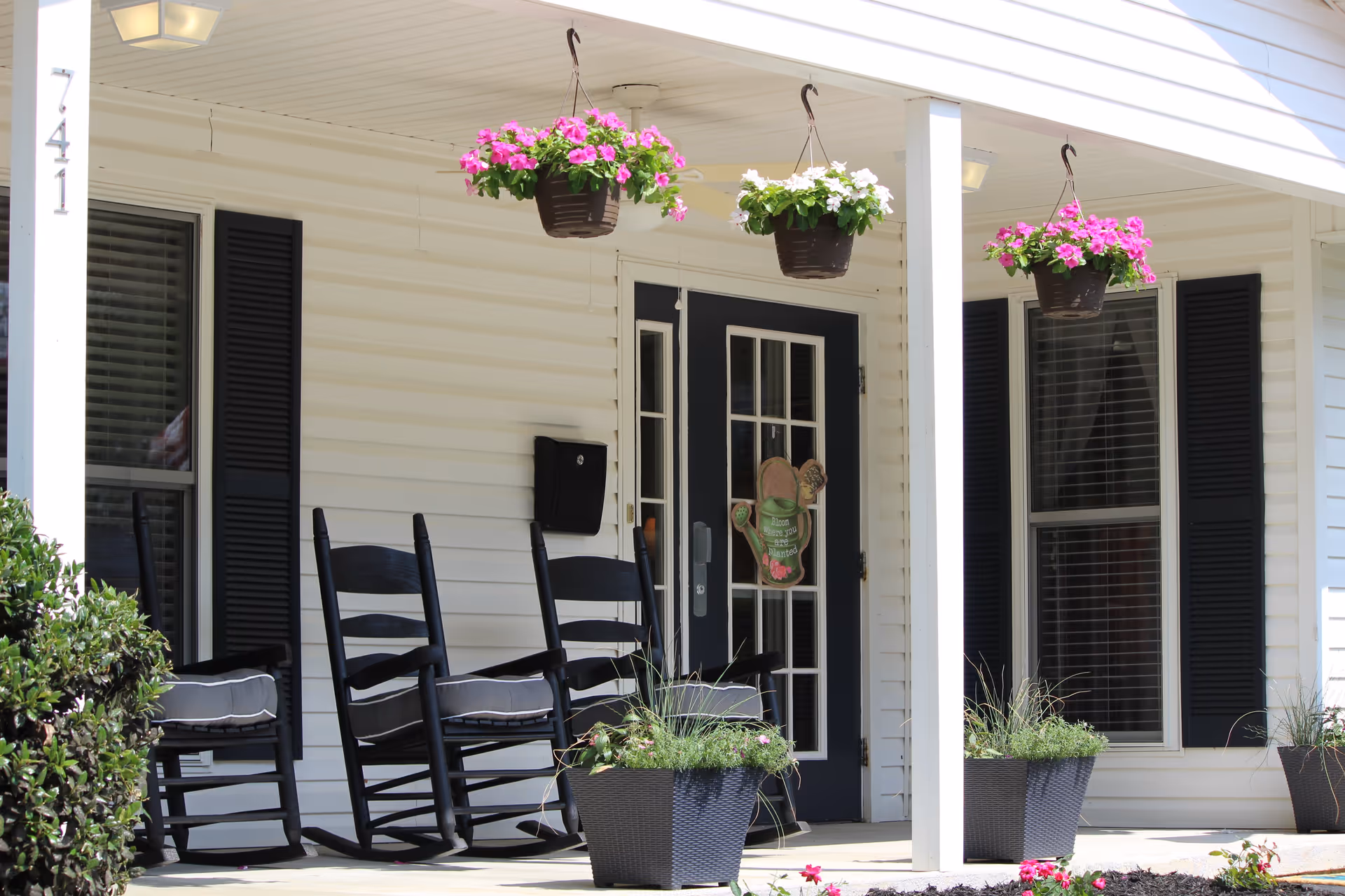 Front porch of a building with white siding and black shutters, featuring three black rocking chairs with cushions, hanging flower baskets with pink and white flowers, potted plants, and a black door with glass panels and a decorative sign.