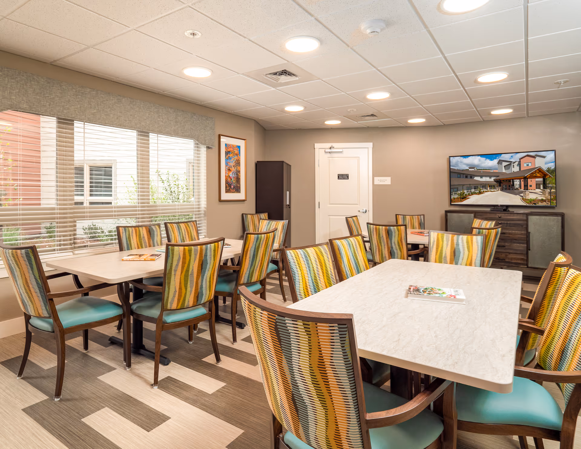 A well-lit dining room in a senior living community with multiple tables and colorful patterned chairs. Large windows with blinds allow natural light to enter. A flat-screen TV mounted on the wall displays an image of the facility's exterior. The room has a modern ceiling with recessed lighting and neutral-toned walls and flooring.