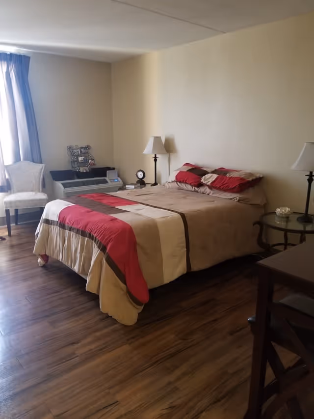 A bedroom with a bed covered in a beige, red, and brown comforter, two matching pillows, two bedside tables each with a lamp, a clock and some books on one table, a chair near a window with curtains, and a wooden floor.