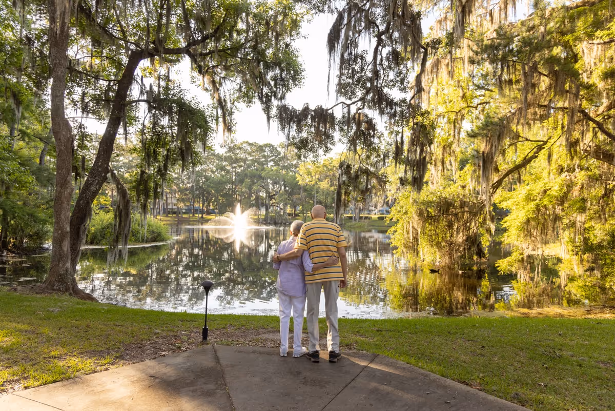 An elderly couple stands arm in arm on a paved path, looking out over a serene pond with a water fountain in the center, surrounded by lush green trees draped with Spanish moss in a peaceful outdoor setting.