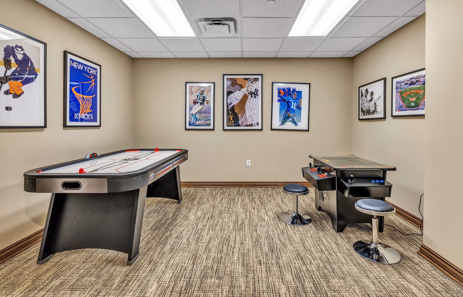 A recreation room with an air hockey table on the left and an arcade game table on the right, accompanied by two black stools. The beige walls are decorated with framed sports-themed artwork, including images related to basketball, football, and baseball. The room has a carpeted floor and a drop ceiling with fluorescent lighting.
