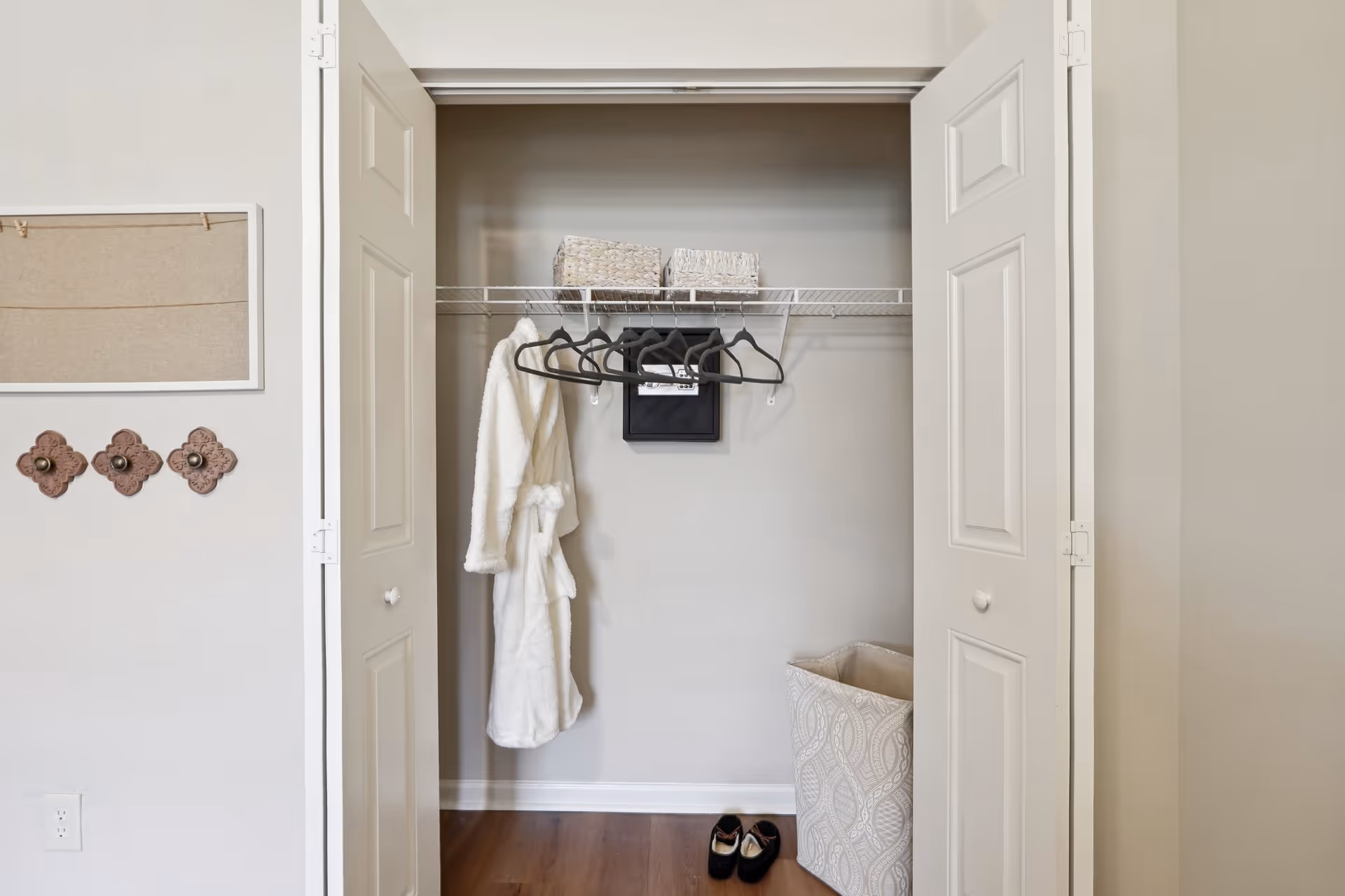 Open closet with white double doors showing a white bathrobe hanging on a black hanger, several empty black hangers on a wire shelf, two woven baskets on the shelf, a patterned laundry hamper on the floor, and a pair of black shoes on the wooden floor. To the left of the closet, there is a wall with three decorative hooks and a bulletin board.