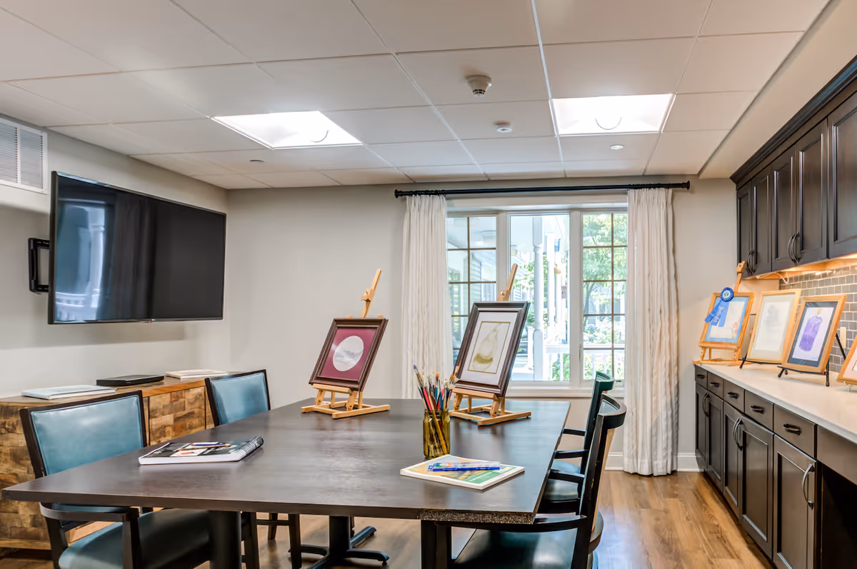 A bright and inviting art room with a large dark table surrounded by chairs. On the table are two small easels holding framed artwork, a jar with paintbrushes, and some art supplies. The room has a large window with white curtains letting in natural light, dark wooden cabinets along one wall displaying more framed artwork, and a flat-screen TV mounted on the opposite wall.