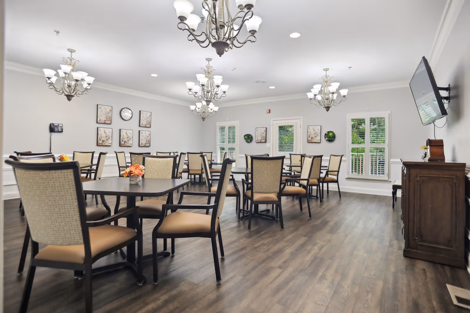 A bright and spacious dining room with multiple tables and chairs arranged neatly. The room features elegant chandeliers hanging from the ceiling, framed artwork on the walls, a clock, and large windows with white shutters allowing natural light to enter. A flat-screen TV is mounted on the right wall above a wooden cabinet.