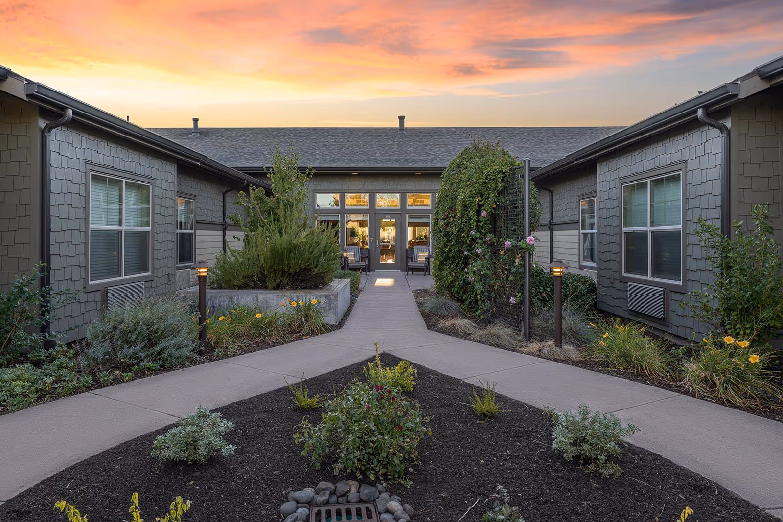 Outdoor courtyard area at sunset with a paved walkway leading to a building entrance. The courtyard is landscaped with bushes, flowers, and small trees on either side of the path. The building has gray siding and multiple windows.