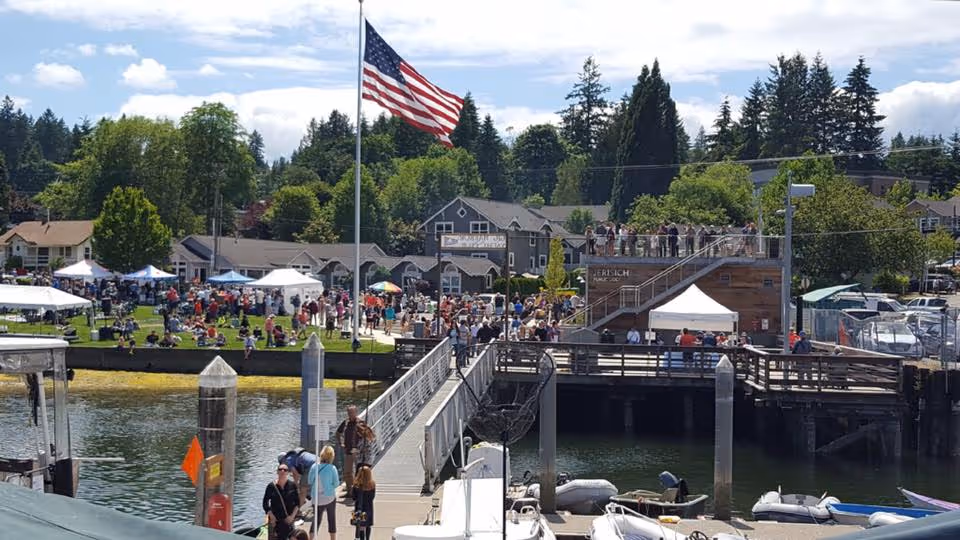 Crowded waterfront park and dock with an American flag, boats, tents, and people gathered for an outdoor event.