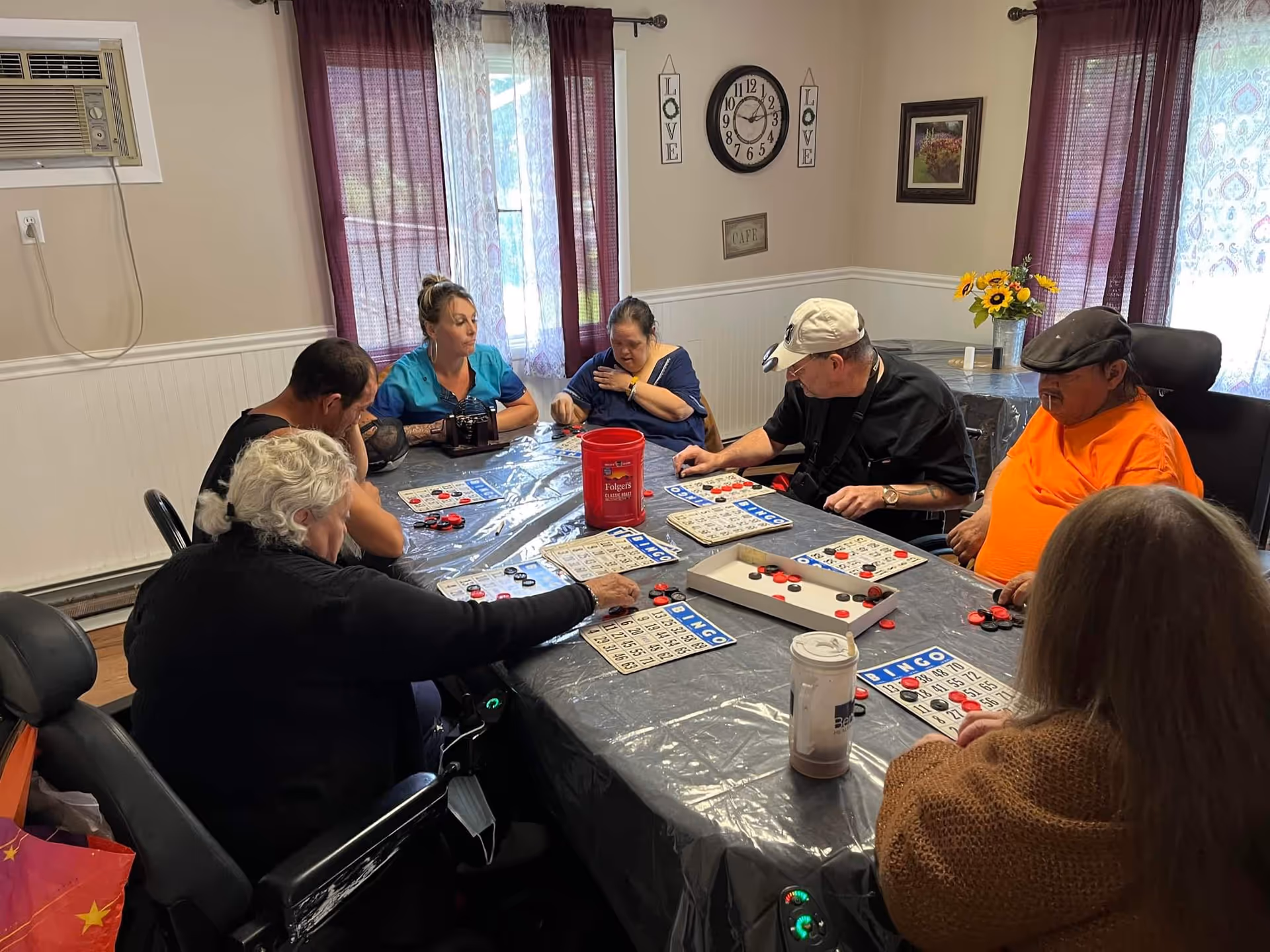 A group of seven people sitting around a table playing bingo in a room with beige walls, purple curtains, and wall decorations including a clock and signs that say 'LOVE'. The table is covered with a plastic tablecloth and has bingo cards, chips, and a red container on it.