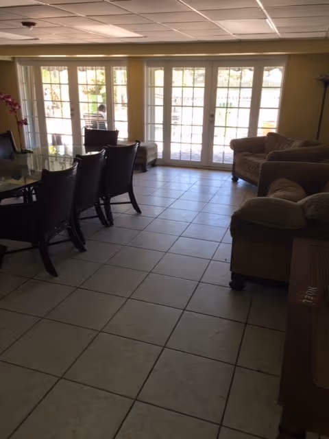 Tile-floored common room with a dining table and chairs on the left, upholstered sofas on the right, and large glass French doors letting in daylight.