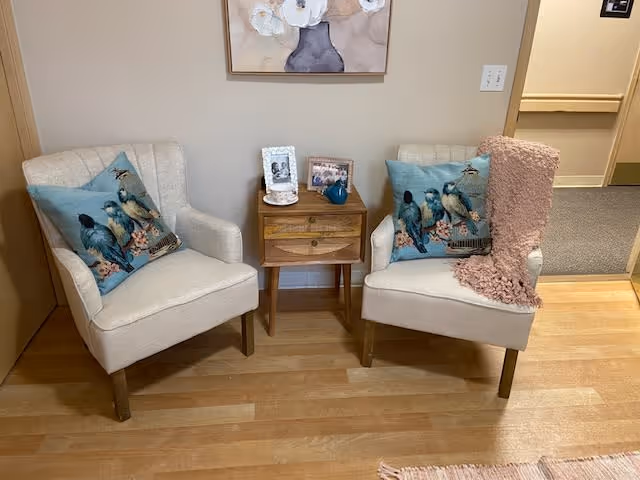 Two beige upholstered armchairs with blue bird-themed pillows are positioned on either side of a small wooden side table. The table holds two framed photos and a small blue decorative item. A pink knitted throw blanket is draped over the right chair. The setting is indoors with light wood flooring and a beige wall with a framed floral painting above the chairs.