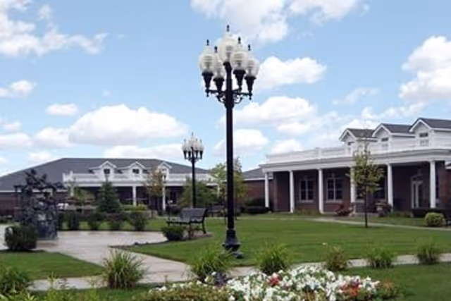Outdoor view of a senior living facility courtyard with green lawns, flower beds, benches, decorative street lamps, and buildings with white trim under a partly cloudy sky.