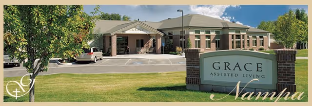 Exterior view of Grace Assisted Living facility in Nampa, showing a single-story brick building with a parking lot and a large sign in front reading 'Grace Assisted Living'. There is a tree and green lawn in the foreground under a partly cloudy sky.
