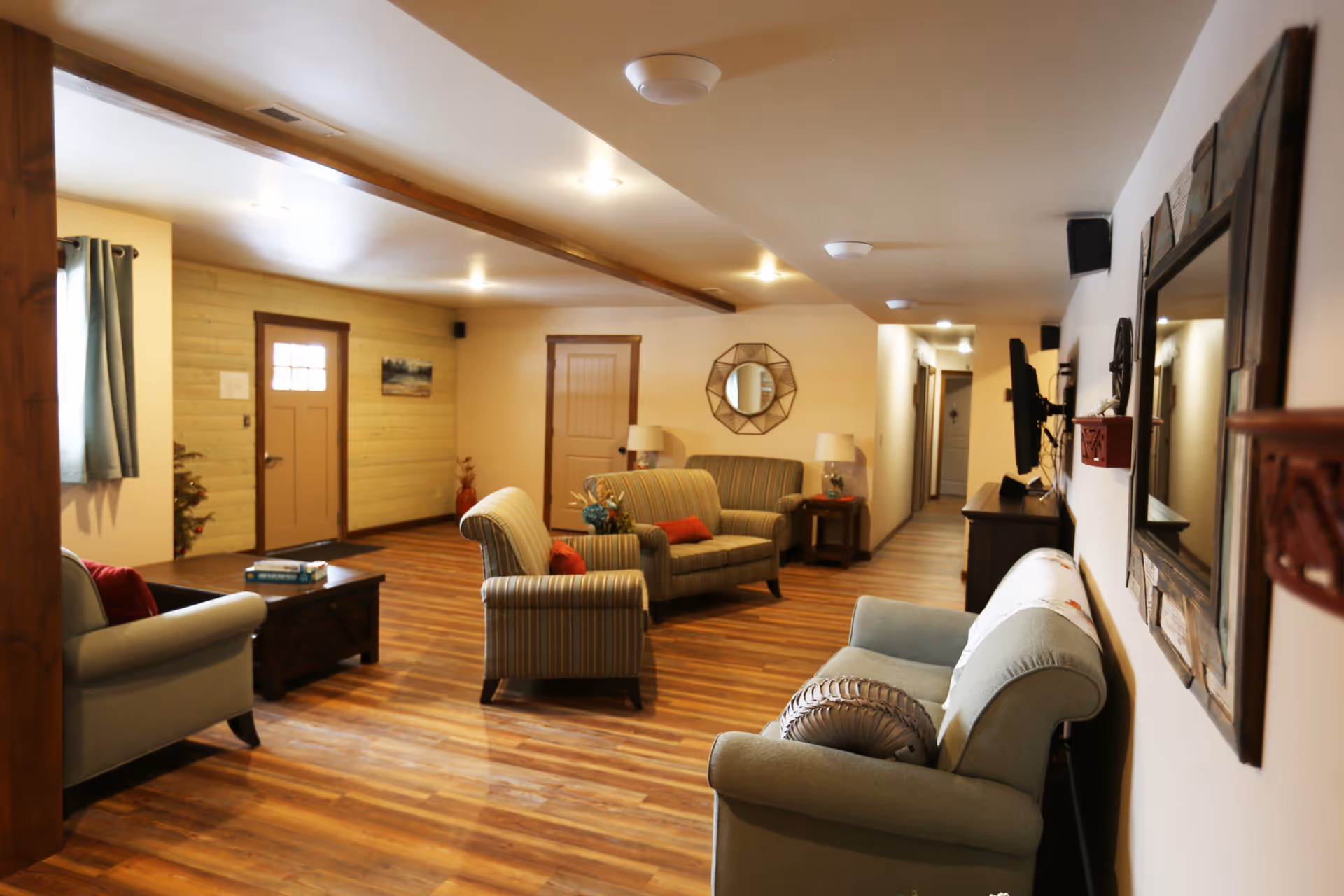 A cozy living room area in Hidden Meadow Memory Care with wooden flooring, several upholstered armchairs and sofas with cushions, a coffee table with books, a wall mirror, lamps on side tables, and a hallway leading to other rooms.