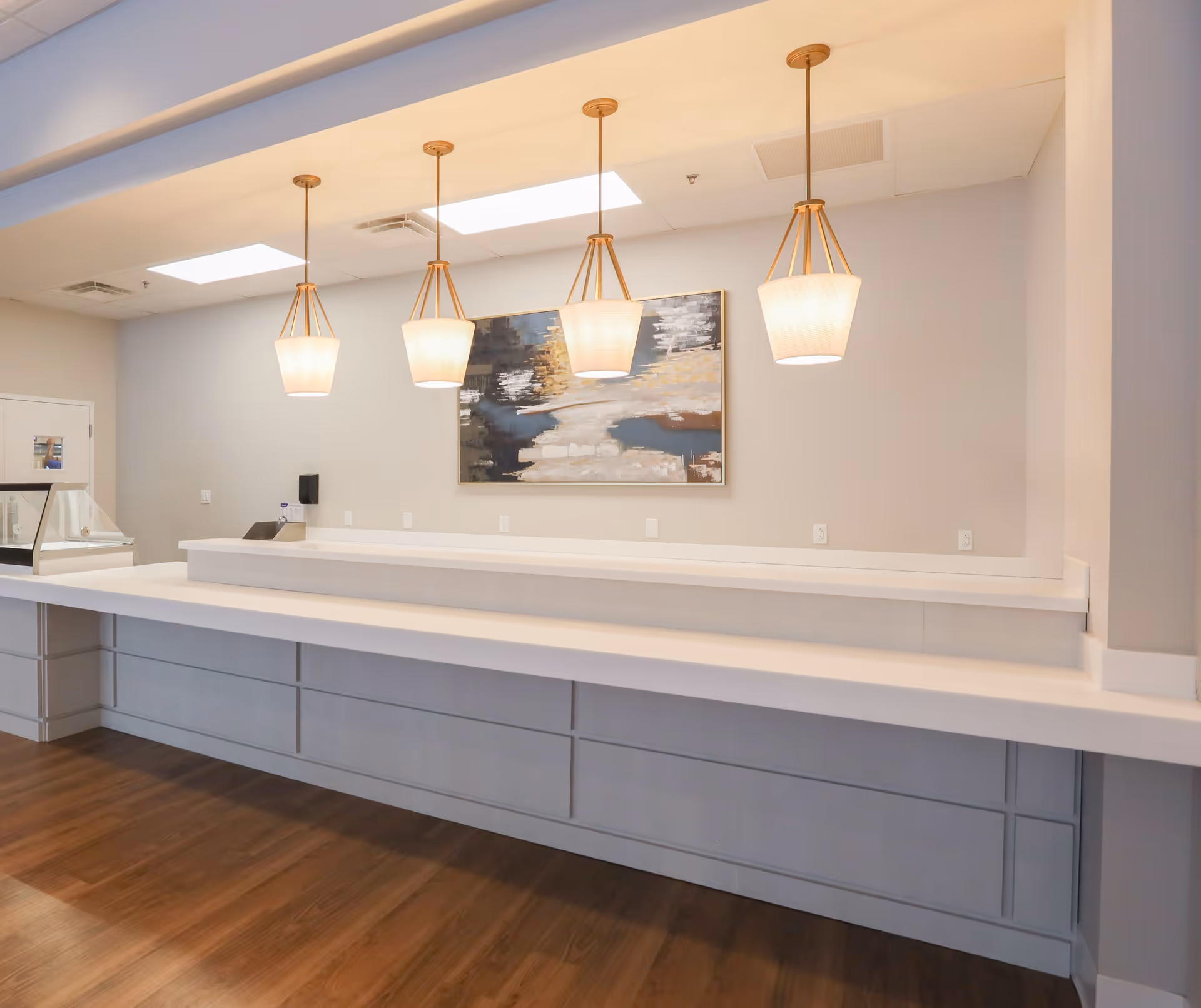 Interior view of a modern serving counter area with four pendant lights hanging from the ceiling, a large abstract painting on the wall, and a wooden floor. The counter is white with a raised section and there are multiple electrical outlets on the wall behind it.