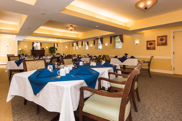 A dining room with multiple tables covered in white tablecloths and blue napkins. Each table is set with cups, saucers, and silverware. The room has yellow walls, carpeted floors, and several windows with curtains. There are framed floral pictures on the walls and ceiling lights providing warm illumination.