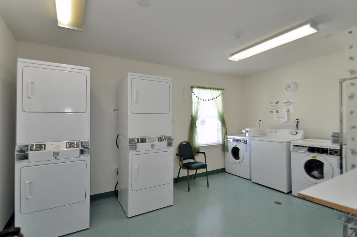 Laundry room with two stacked washer and dryer units on the left, three front-loading washing machines on the right, a green chair near a window with green curtains, and a clock on the wall.