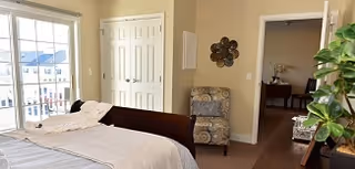 A bright bedroom in a senior living community featuring a bed with white linens, a cushioned armchair with a decorative pillow, a wall-mounted metal art piece, a wooden dresser with a green plant, and a doorway leading to another room. Large windows allow natural light to fill the space.