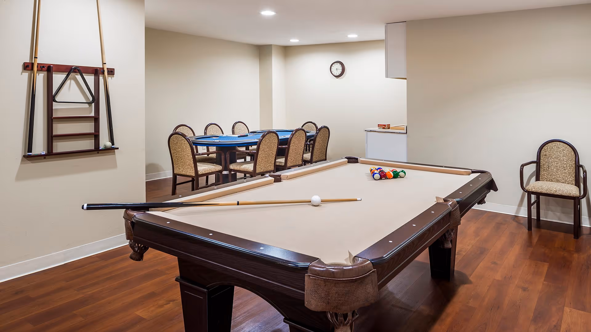 A game room with a beige pool table in the foreground, pool cues resting on the table, and a rack of cues mounted on the wall. In the background, there is a table with several chairs around it, a clock on the wall, and a small counter area. The room has wooden flooring and beige walls.