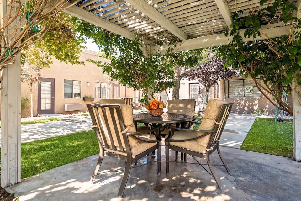 Outdoor seating area under a white pergola with green vines and foliage providing shade. There is a round metal table with a flower arrangement in the center and six cushioned chairs around it. The area is surrounded by a concrete pathway, green grass, and beige building walls with windows and doors in the background.