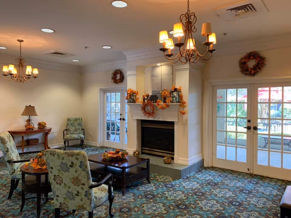 A cozy living room area with floral patterned armchairs arranged around a dark wooden coffee table. The room features a fireplace decorated with autumn-themed wreaths and garlands. Two chandeliers hang from the ceiling, and there are double glass doors leading outside, allowing natural light to enter. The carpet has a blue and brown intricate pattern, and a side table with a lamp and small decorations is visible in the corner.