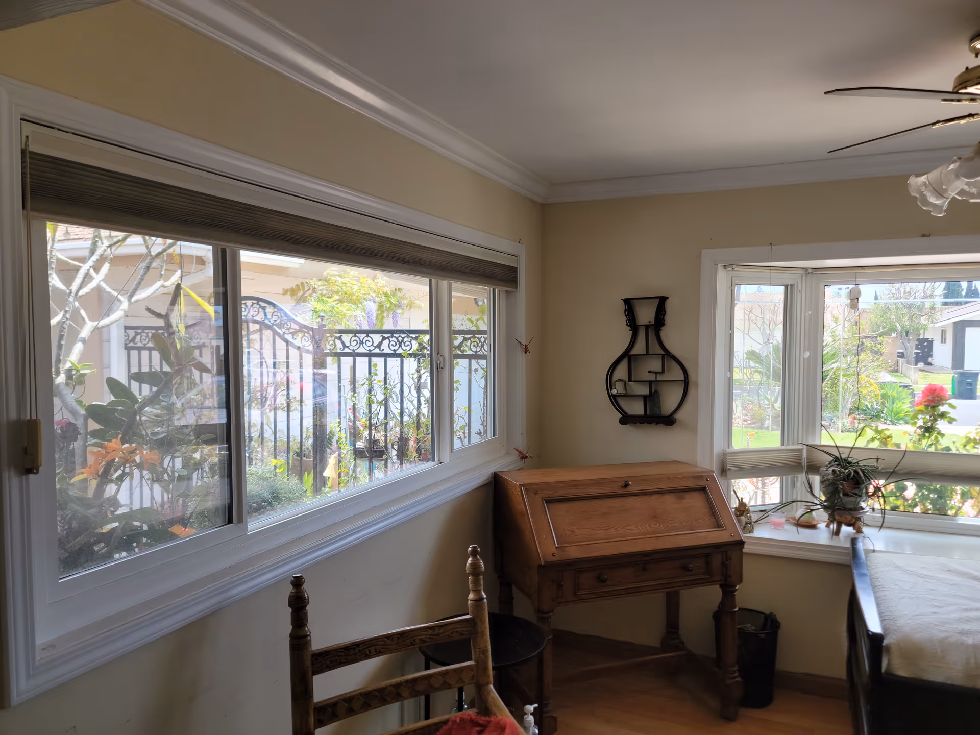 Sunlit dining area with large windows overlooking a garden, featuring a wooden roll-top desk, chairs, and plants on the window sill.