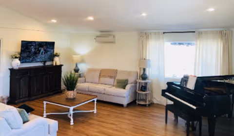 A bright living room with wooden flooring featuring a beige sofa with cushions, a white coffee table with a plant on it, a dark wooden TV stand with a flat-screen TV and decorative items, a black grand piano with sheet music, and two lamps near the sofa and window with white curtains.