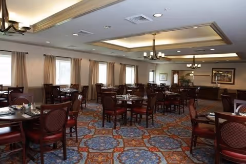 A spacious dining room with multiple dark wood tables and chairs arranged neatly. The room has large windows with beige curtains allowing natural light to enter. The ceiling features recessed lighting and decorative chandeliers. The carpet has a patterned design in blue, orange, and beige tones. There are framed pictures and plants along the walls.