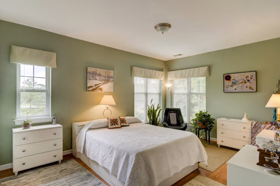 A bedroom with light green walls featuring a large bed with a white quilt and two decorative pillows. There are two white dressers on either side of the room, a black chair near the windows, several plants, and framed artwork on the walls. The room is well-lit with natural light coming through the windows and lamps providing additional lighting.