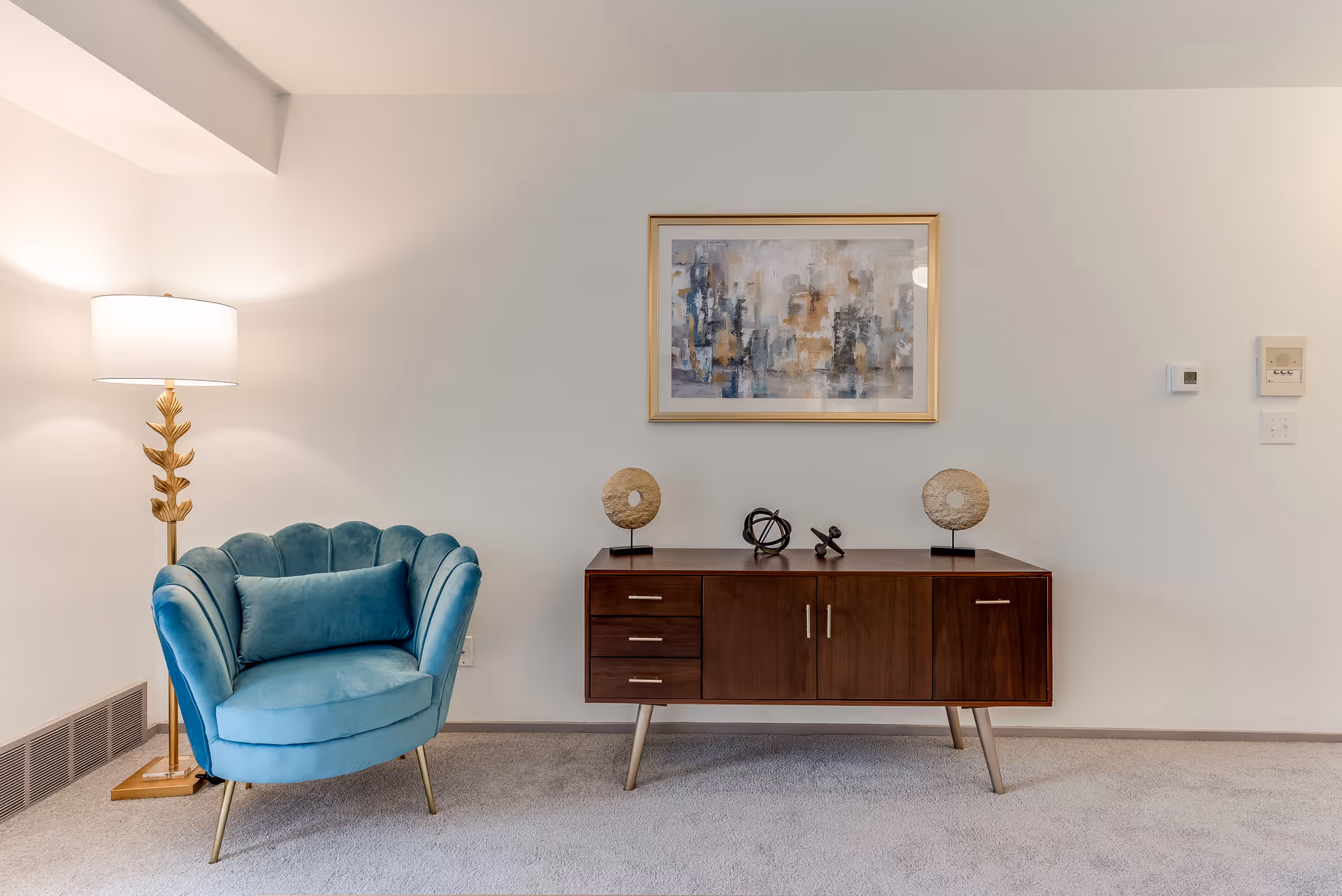A cozy corner of a room featuring a teal upholstered armchair with a matching cushion, a tall floor lamp with a gold leaf design, and a mid-century modern wooden sideboard with decorative items on top. A framed abstract painting hangs on the wall above the sideboard.