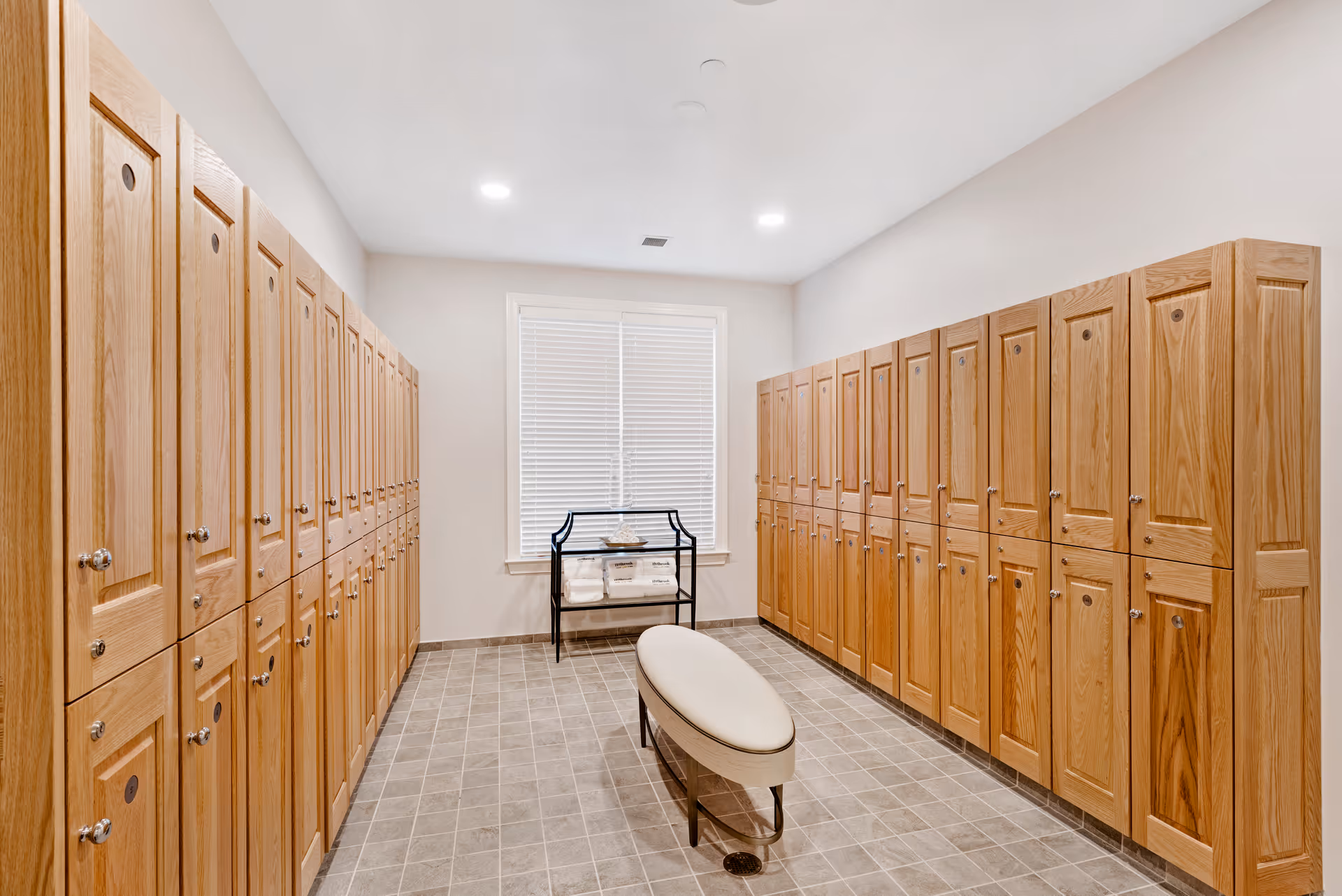A locker room with two rows of wooden lockers on either side, a cushioned bench in the center, and a window with blinds at the far end. A small black metal rack with folded towels is placed under the window. The room has tiled flooring and recessed ceiling lights.