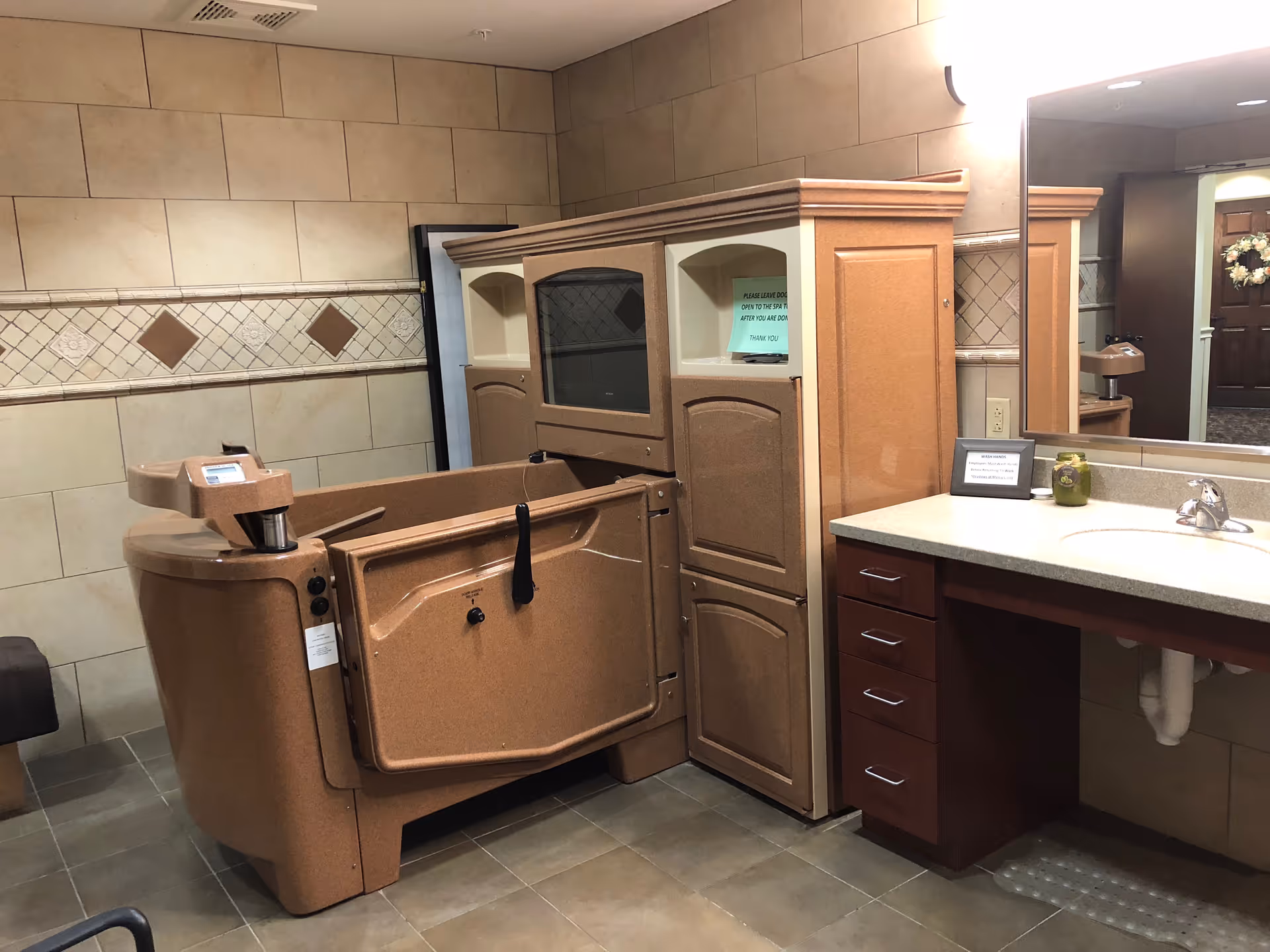A bathroom with beige tiled walls and floor, featuring a large accessible bathtub with a door for easy entry. There is a countertop with a sink and a mirror above it. The room has a warm, neutral color palette and appears clean and well-maintained.