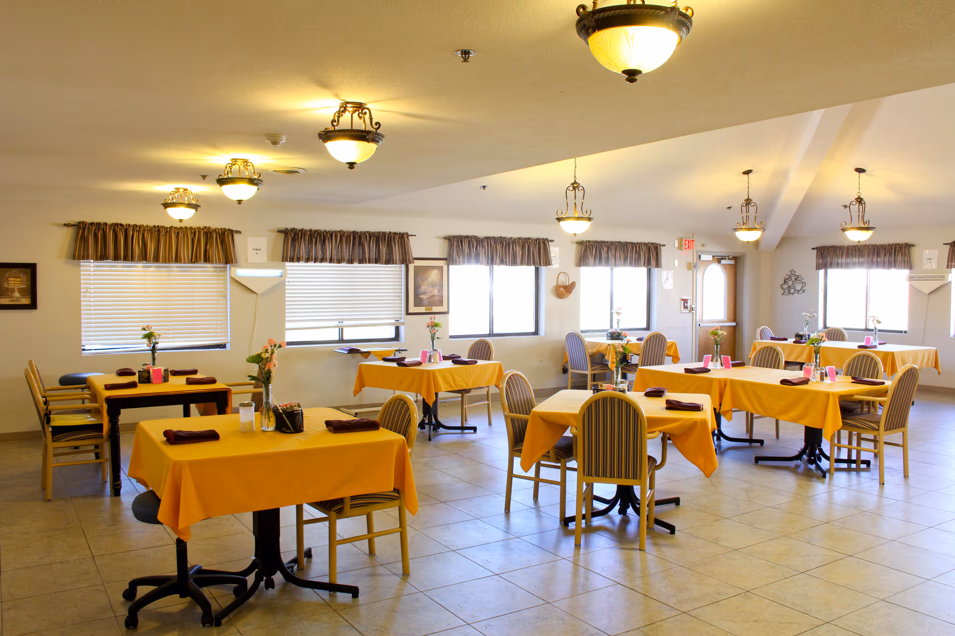 A bright dining room with multiple tables covered in yellow tablecloths, each set with napkins and small flower vases. The room has several windows with brown valances, tiled floors, and ceiling lights providing warm illumination.
