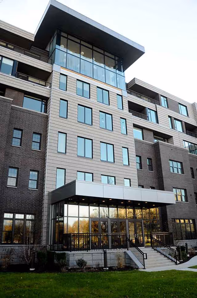 Modern multi-story senior living building showing the front entrance with a large glass lobby, steps, and upper-floor balconies.