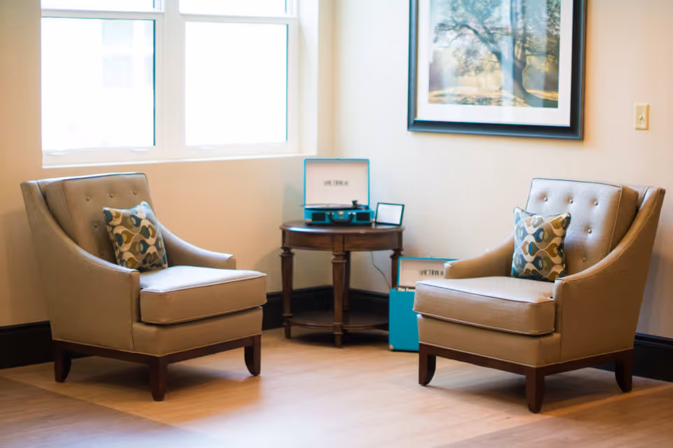 Two beige tufted armchairs with patterned pillows face each other around a small round table holding a teal record player in a bright seating area with a window and framed art.