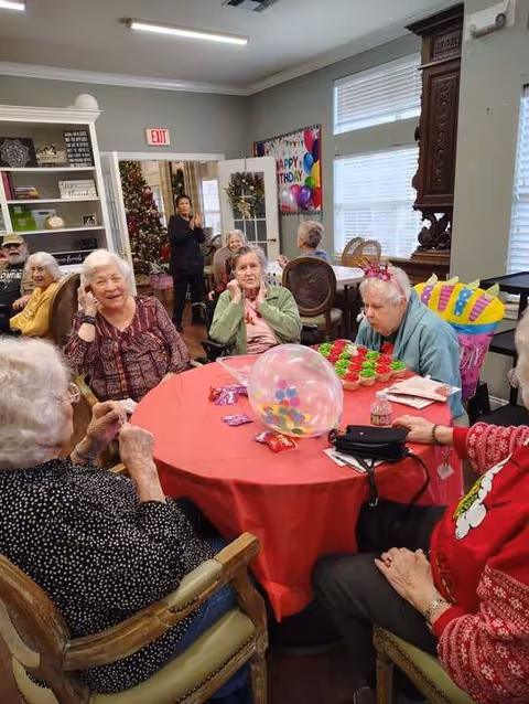 A group of elderly people sitting around a table with a red tablecloth in a decorated room. There are colorful balloons, a birthday banner, and cupcakes on the table. A Christmas tree is visible in the background along with a woman standing near the door.
