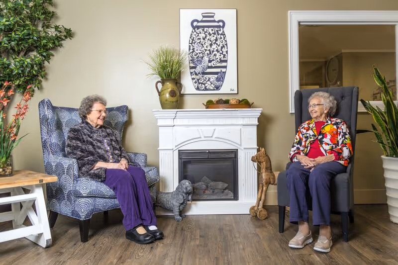 Two elderly women sitting in armchairs facing each other in a cozy room with a white decorative fireplace between them. The room has wooden flooring, plants, and a framed artwork above the fireplace. Both women are smiling and appear to be engaged in conversation.