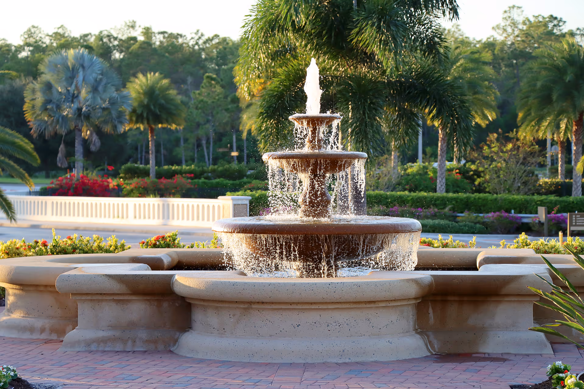 A multi-tiered stone water fountain with water cascading down each level, surrounded by a circular stone bench. In the background, there are palm trees, bushes, and a white railing with a road and forested area beyond.