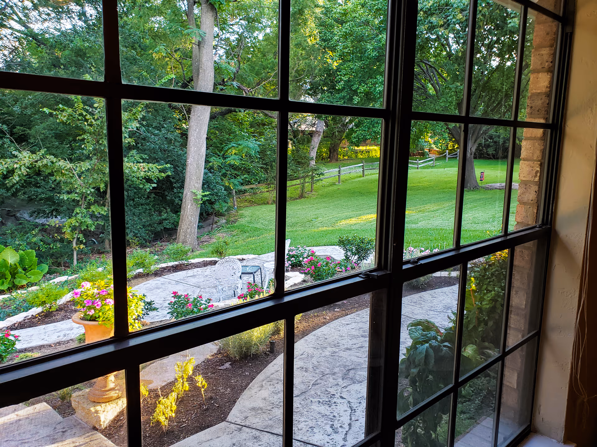 View through a large window with black grid framing, showing a landscaped garden with a curved stone pathway, flower beds with colorful flowers, green grass, trees, and a white metal chair and table set on a stone patio.
