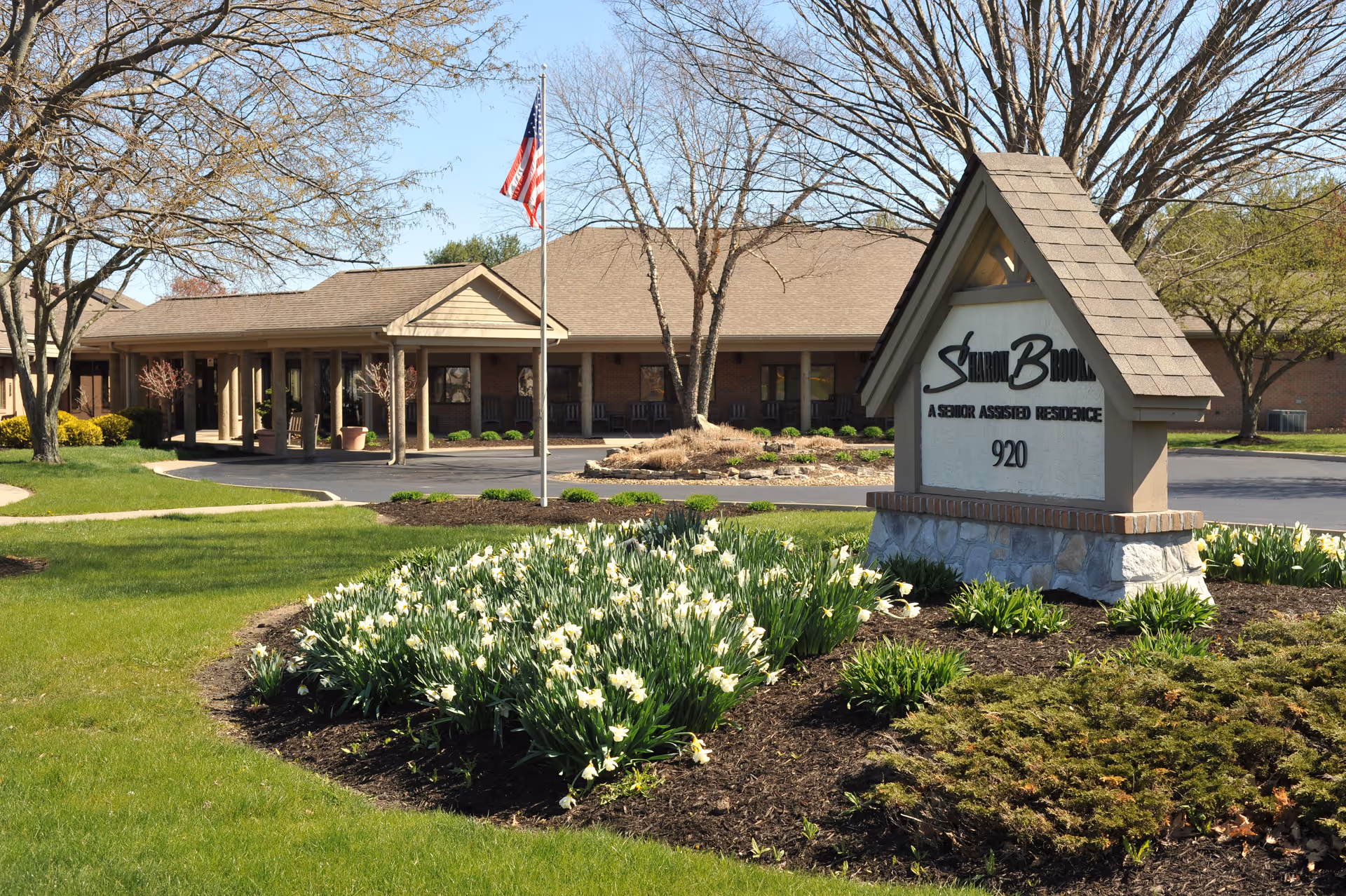 Exterior view of SharonBrooke Assisted Living facility showing a single-story building with a covered entrance, an American flag on a flagpole, and a landscaped garden with blooming white flowers and green grass in the foreground. A large sign with the facility name and address number 920 is prominently displayed near the entrance.