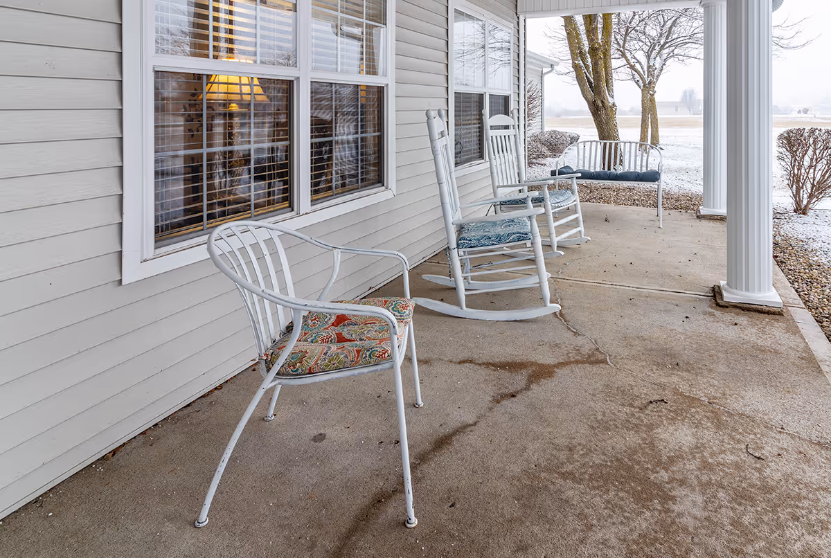 Covered front porch with white rocking chairs, a metal chair with a patterned cushion, and a bench beside windows.
