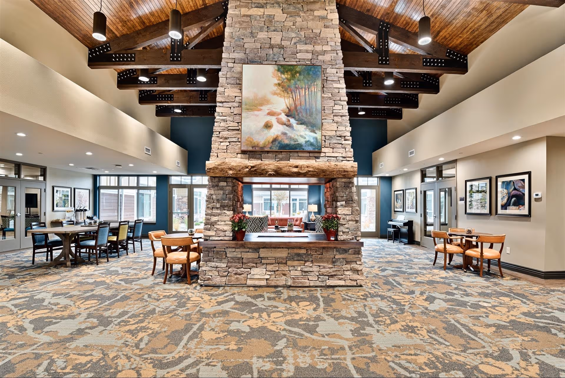 Spacious senior living facility common area with a large stone fireplace in the center, wooden ceiling beams with hanging lights, patterned carpet, multiple seating areas with tables and chairs, large windows allowing natural light, and framed artwork on the walls.