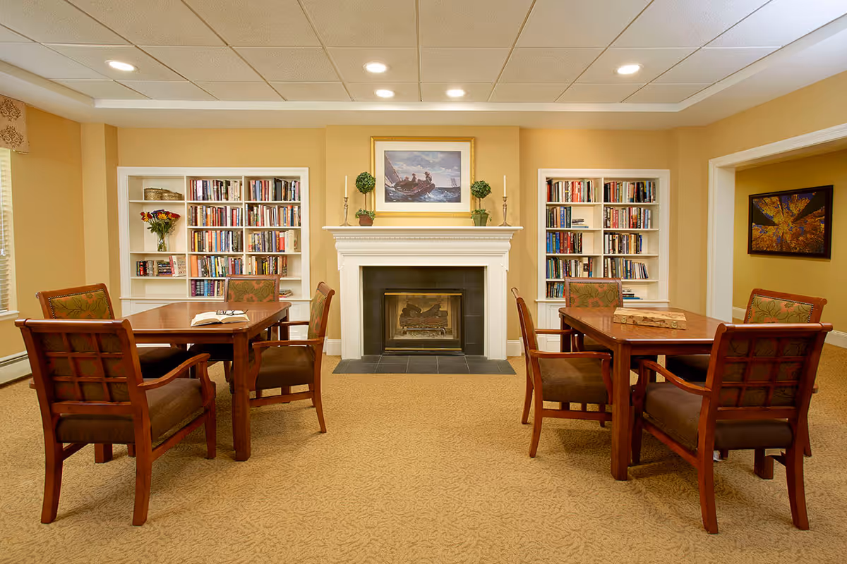 Well-lit common room with two wooden tables and chairs, bookshelves and a fireplace.
