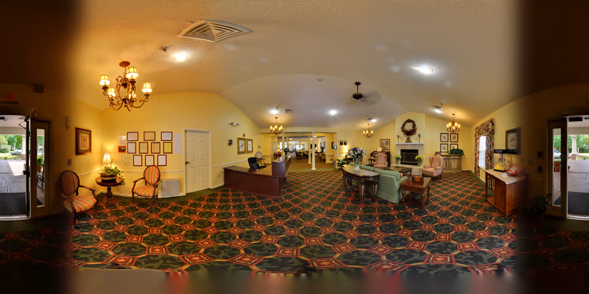 A spacious senior living facility common area with patterned carpet, yellow walls, and multiple seating arrangements including armchairs and sofas. There is a reception desk in the middle, chandeliers hanging from the ceiling, a fireplace with a wreath above it, framed pictures on the walls, and large windows with floral curtains. Double glass doors lead outside on both sides of the room.