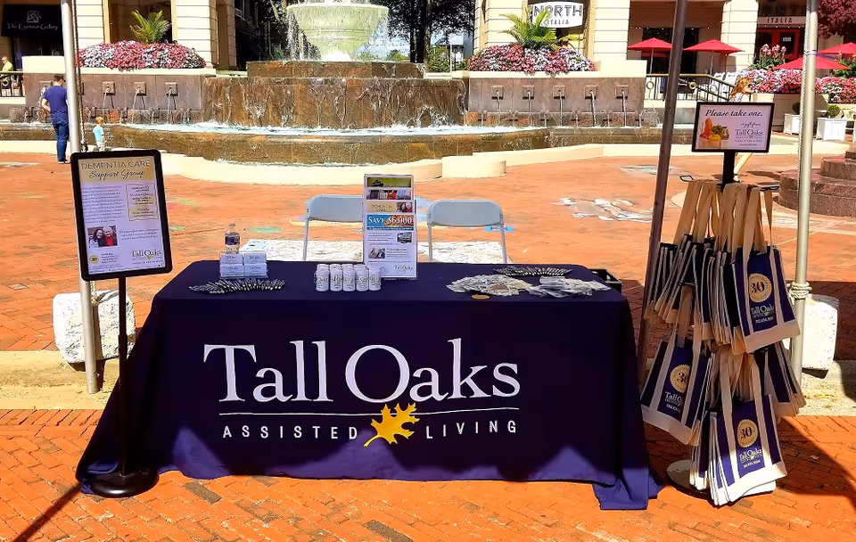 Outdoor promotional table for Tall Oaks Assisted Living set up in front of a large water fountain. The table is covered with a navy blue tablecloth featuring the Tall Oaks logo and has informational pamphlets, small bottles, and pens on it. To the right, there is a stand with multiple Tall Oaks branded tote bags. A sign on the left side of the table mentions a Dementia Care Support Group, and a sign on the right invites people to take a tote bag. The background shows a plaza area with brick flooring, flower beds, and people walking.