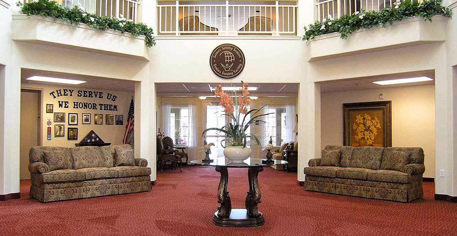 Interior view of a senior living facility lobby with two patterned sofas on either side, a round glass table with a floral arrangement in the center, red carpet flooring, and a wall display honoring veterans with the text 'THEY SERVE US WE HONOR THEM'.