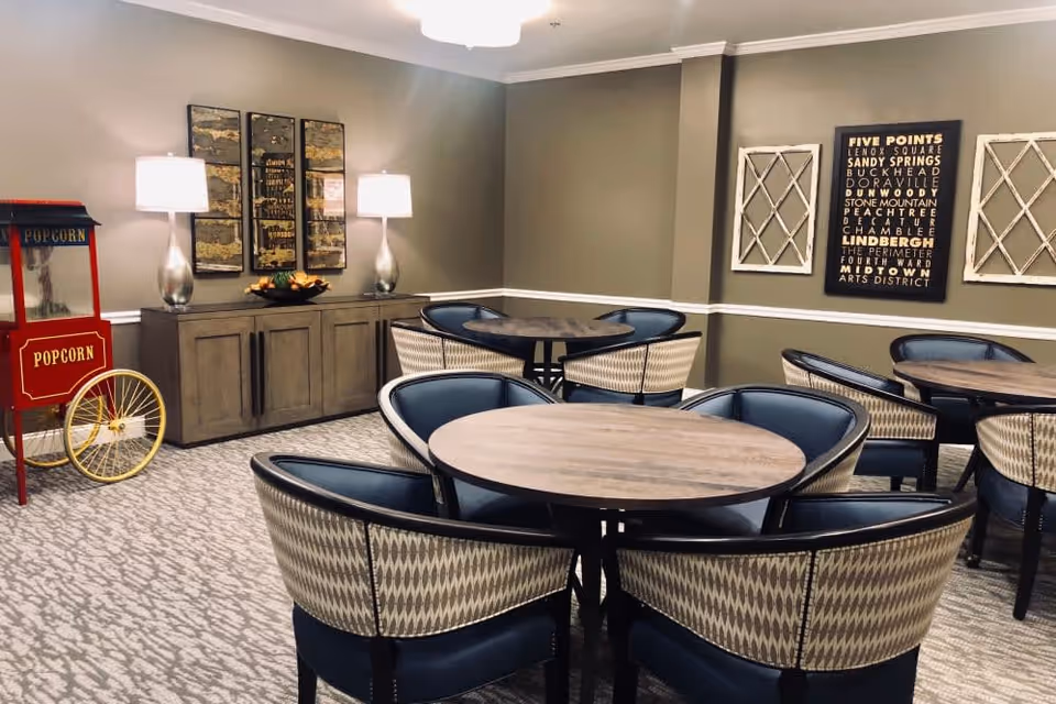 A cozy dining area with round wooden tables surrounded by cushioned chairs with patterned backs. The room has beige walls with white trim, two table lamps on a wooden sideboard, decorative wall art, and a vintage-style red popcorn machine in the corner.