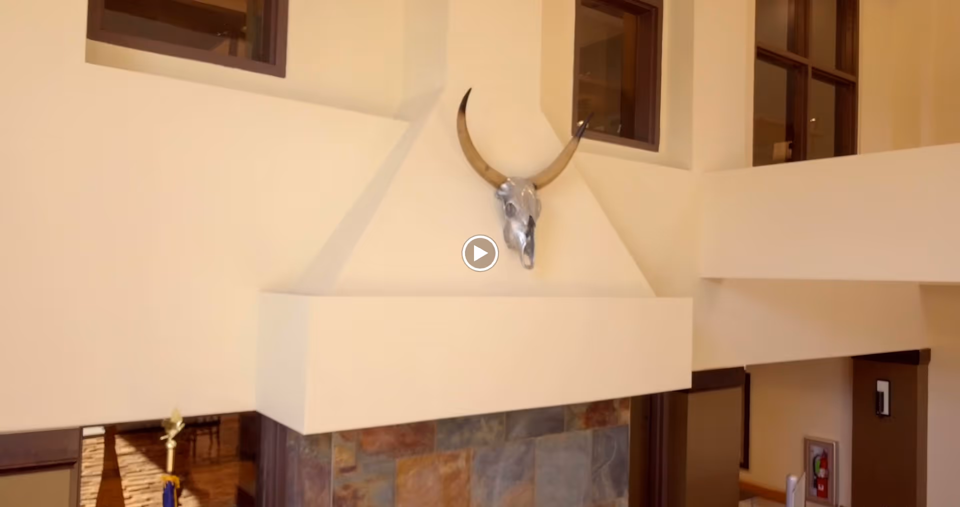 Interior view of a room with a high ceiling featuring a white fireplace with a decorative bull skull mounted above it. The walls are light-colored, and there are several windows near the ceiling allowing natural light in.