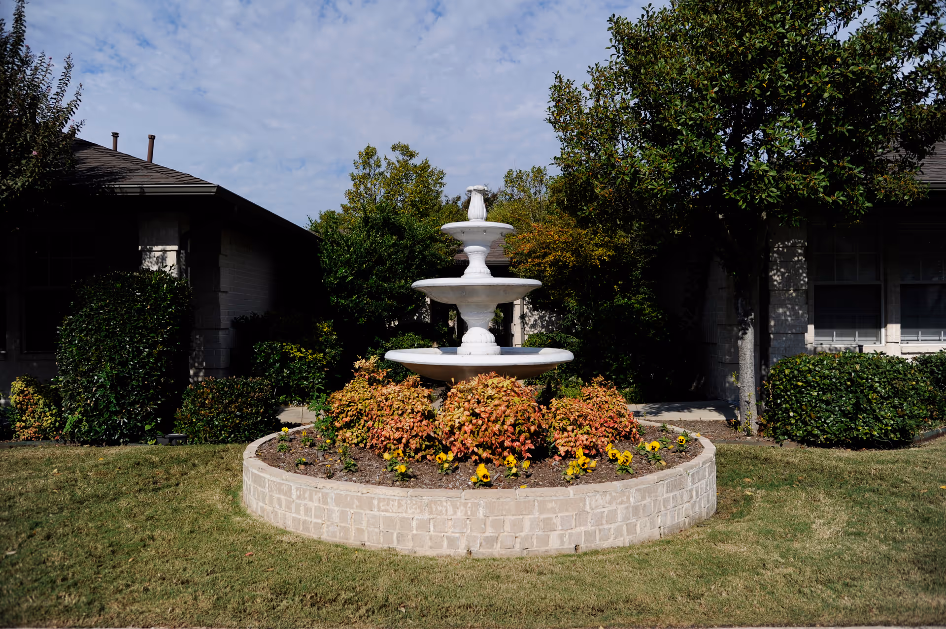 A white three-tiered water fountain surrounded by a circular brick planter filled with green and reddish plants and yellow flowers, set in a grassy area with bushes and trees in front of a building under a partly cloudy sky.