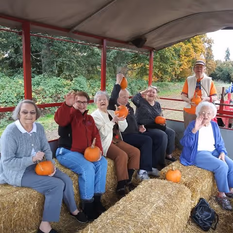 A group of six elderly people sitting on hay bales in an open wagon, each holding small pumpkins and waving. They are outdoors with trees and greenery in the background, enjoying a fall-themed outing.