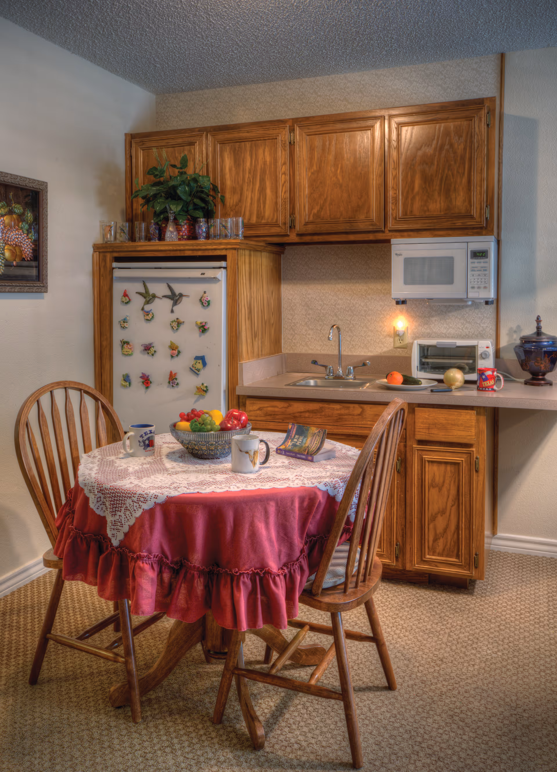 A small kitchen area with wooden cabinets, a white microwave mounted above the counter, a sink, and a refrigerator decorated with colorful magnets. In front of the kitchen is a round wooden table covered with a red tablecloth and a white lace overlay, surrounded by two wooden chairs. On the table are two mugs, a bowl of fruit, and a small book or pamphlet. The floor is carpeted and there is a framed fruit painting on the wall.