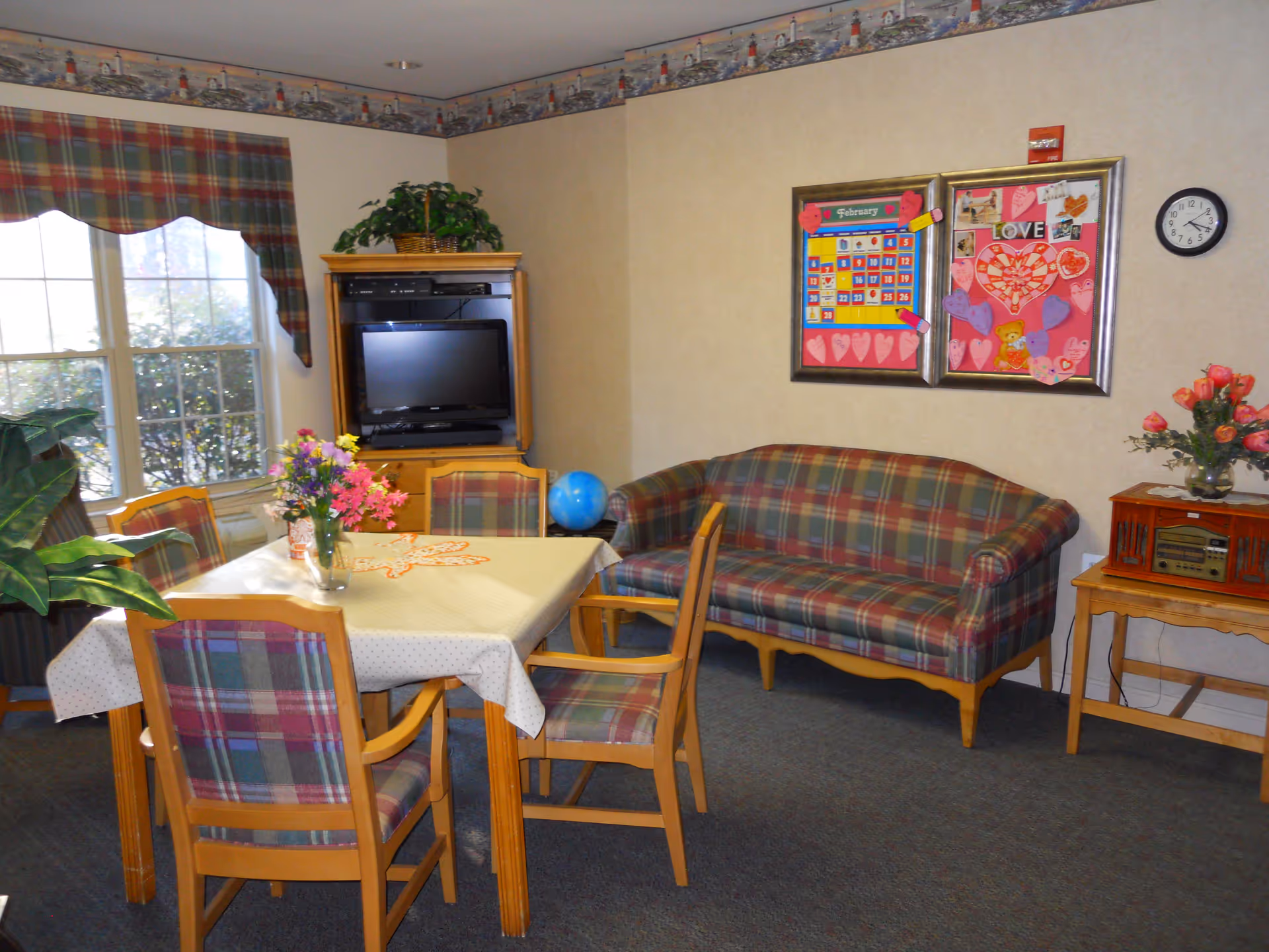 Communal sitting and dining area with a plaid sofa and matching chairs around a table, TV cabinet, and decorated bulletin board on the wall.